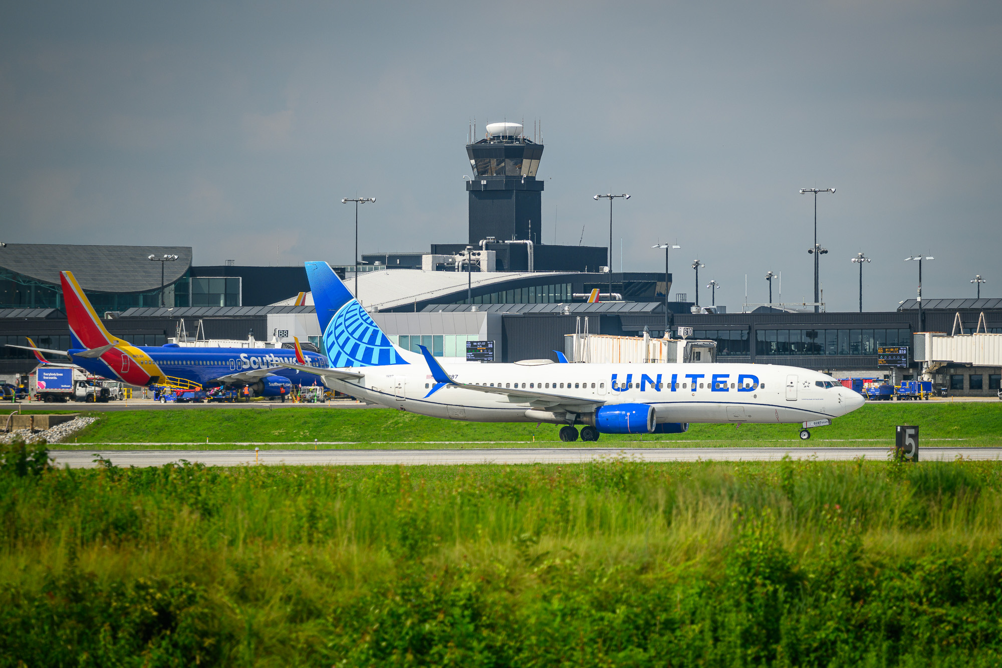 A plane lands on the runway with an airplane and and control tower in the background.