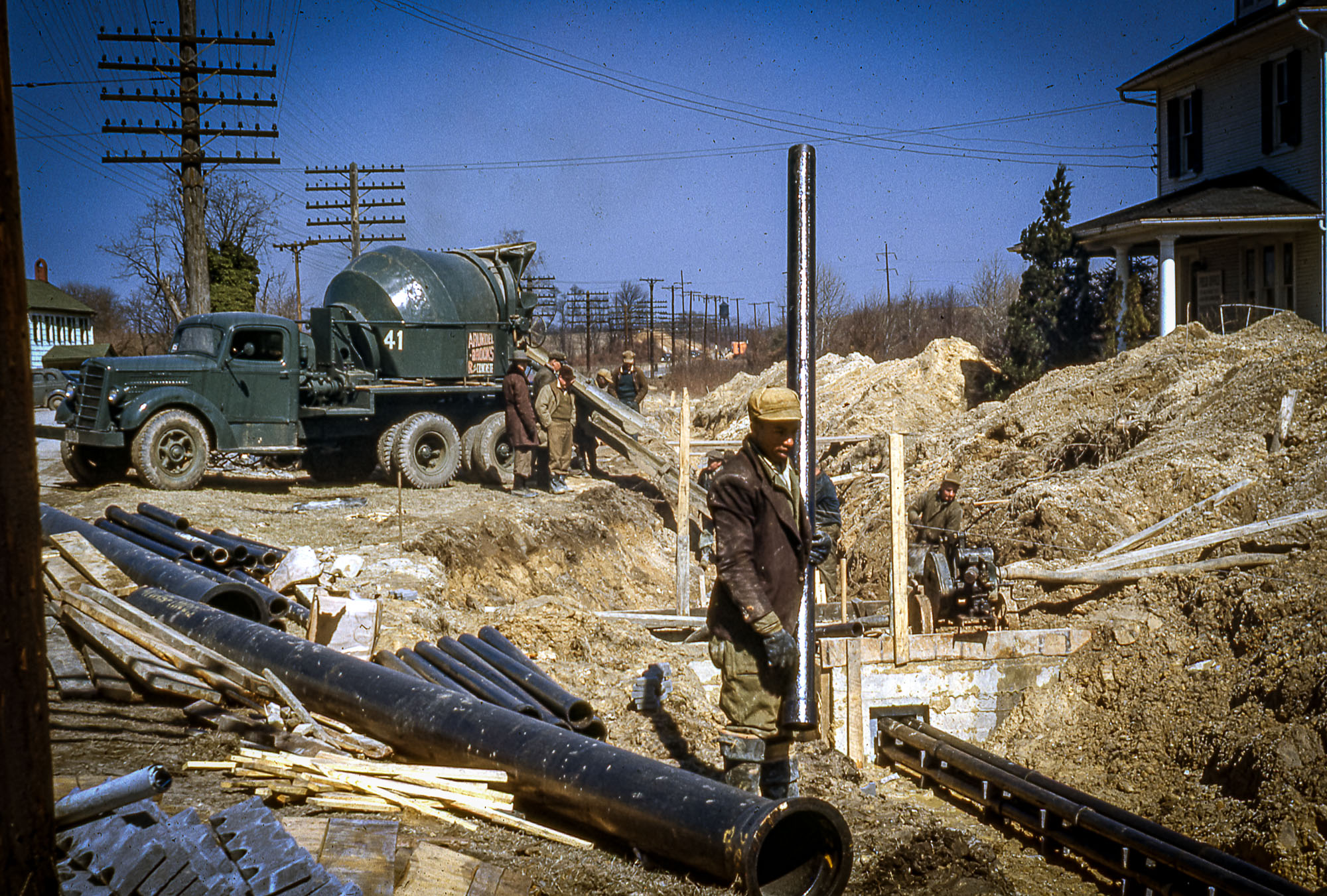 A worker holds a pipe near a trench with a cement mixer and building in the background.