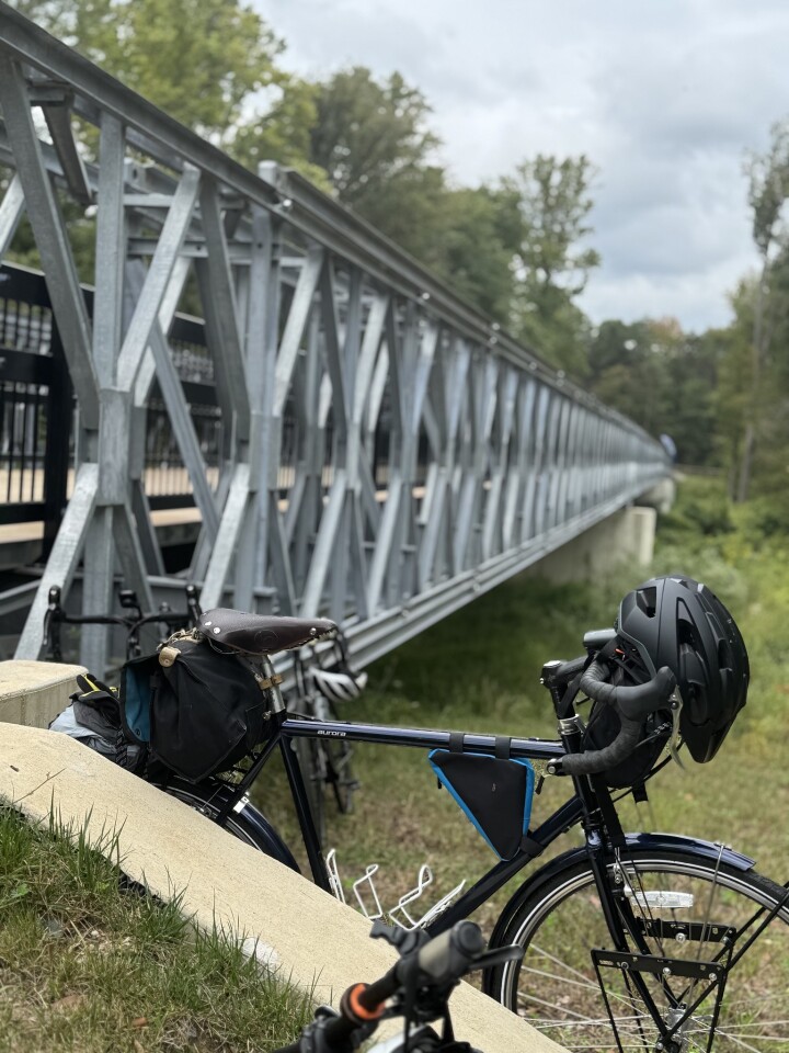 A dark blue touring bicycle with a black helmet and bags parked near a modern, silver metal truss bridge on a cloudy day. Credit: Antoine RJ Wright.