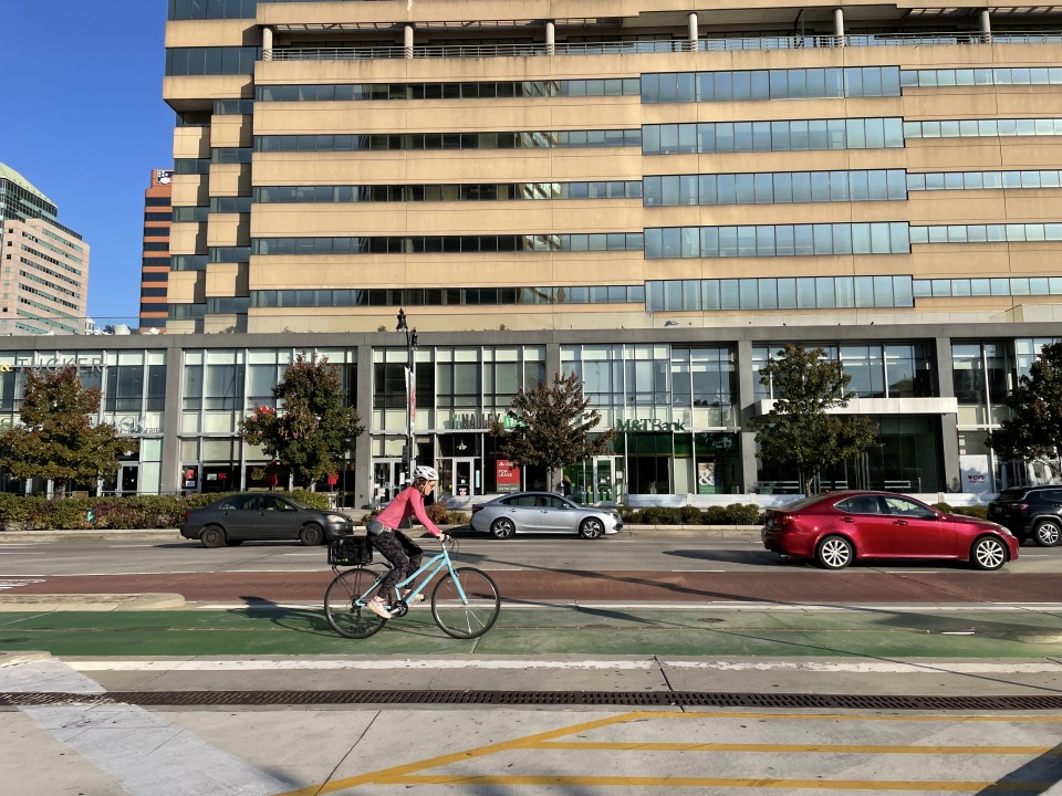 A cyclist on a light blue bike rides along a bright green painted bike lane in a city, with large glass buildings and parked cars in the background.
