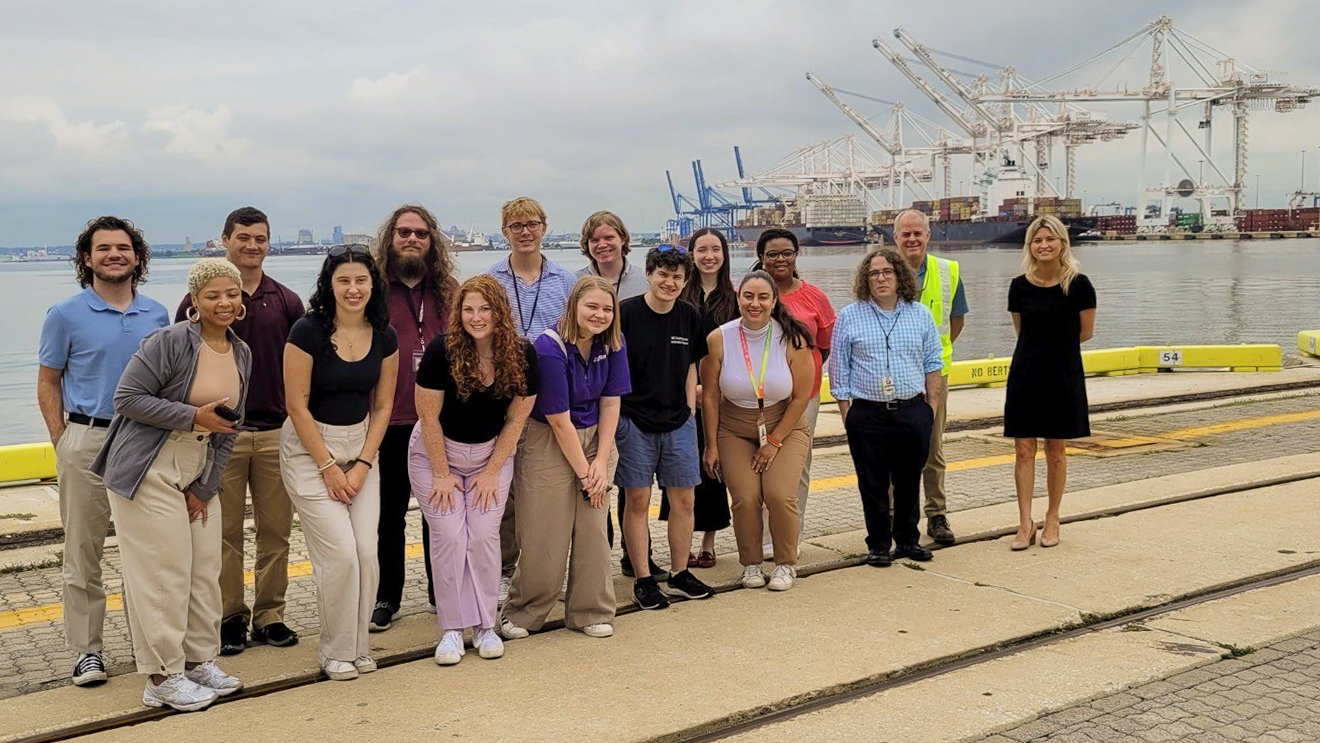 A diverse group of MDOT Fellows and program leaders standing together outside at the Port of Baltimore, with massive shipping cranes behind them.