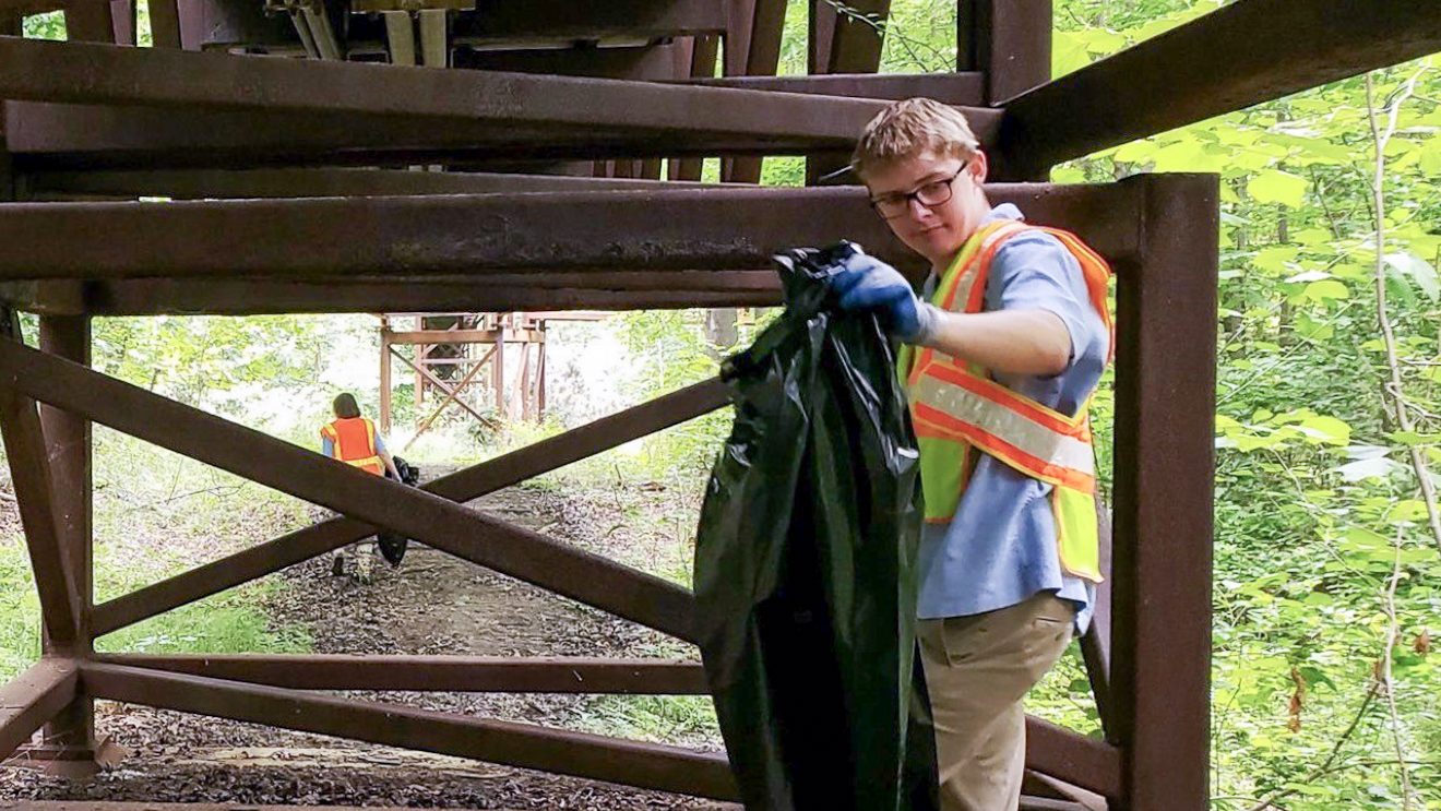 An MDOT Fellow wearing a high-visibility safety vest and gloves smiles while holding open a black trash bag during a community cleanup day.